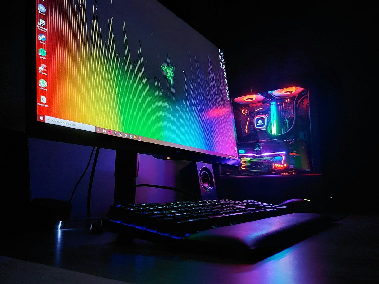 A desk with a wide monitor showing a rainbow spectrum, a keyboard and an RGB-lit tower