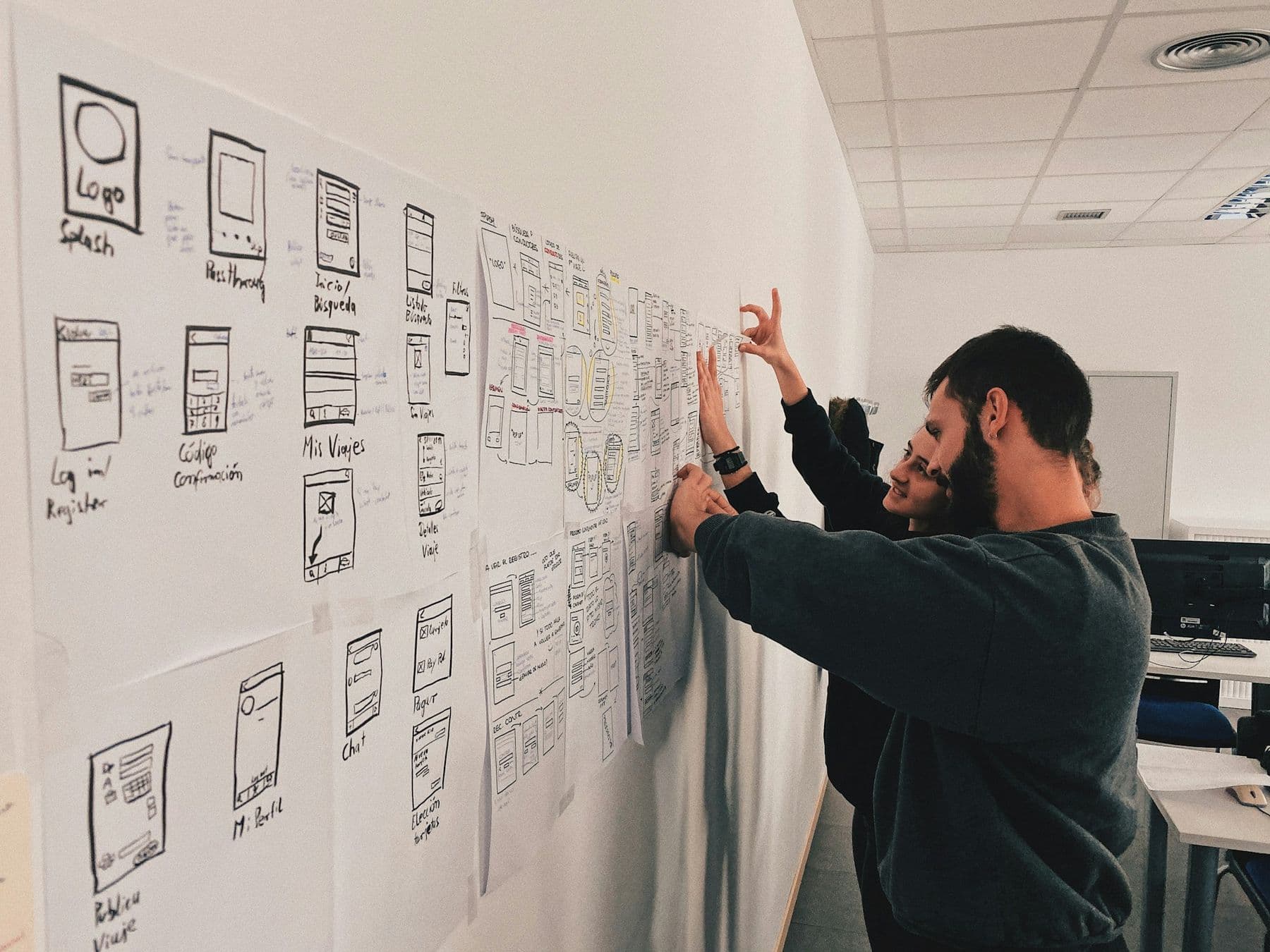 Two people pinning hand-drawn app wireframes to a long white wall